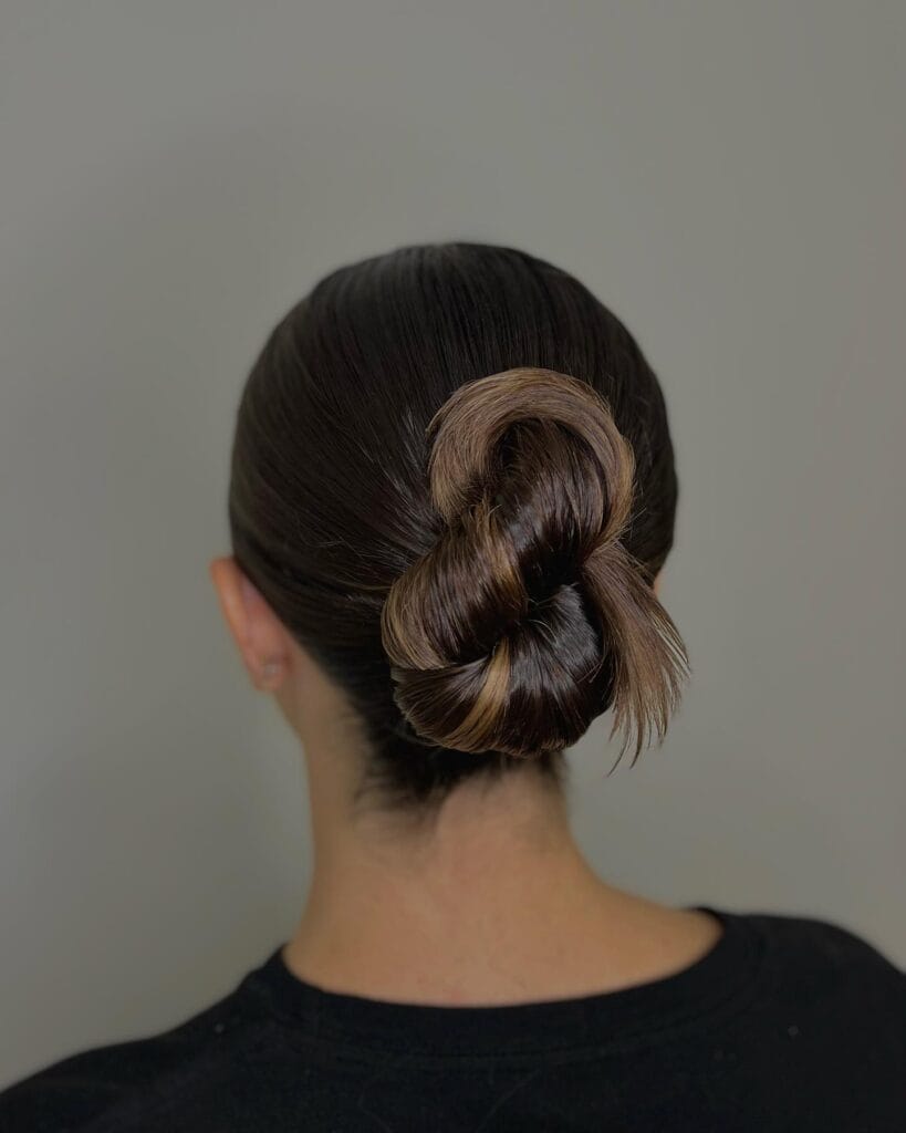 A woman with sleek dark brown hair styled in a modern sculpted low knot with glossy loops.