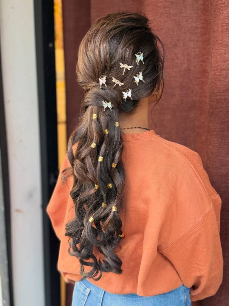 A rear view of a woman with long, dark, curly hair in a low ponytail decorated with gold cuffs and pastel butterfly clips.