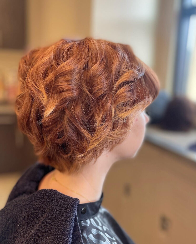 A side view of a woman with a short layered hairstyle featuring thick copper-red curls and a soft nape.