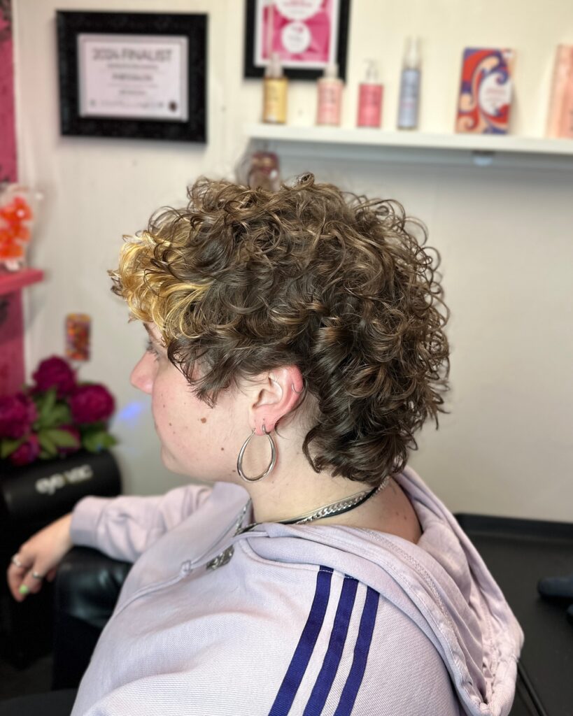 A side view of a woman with a short curly brown hairstyle featuring a bright blonde section at the front.