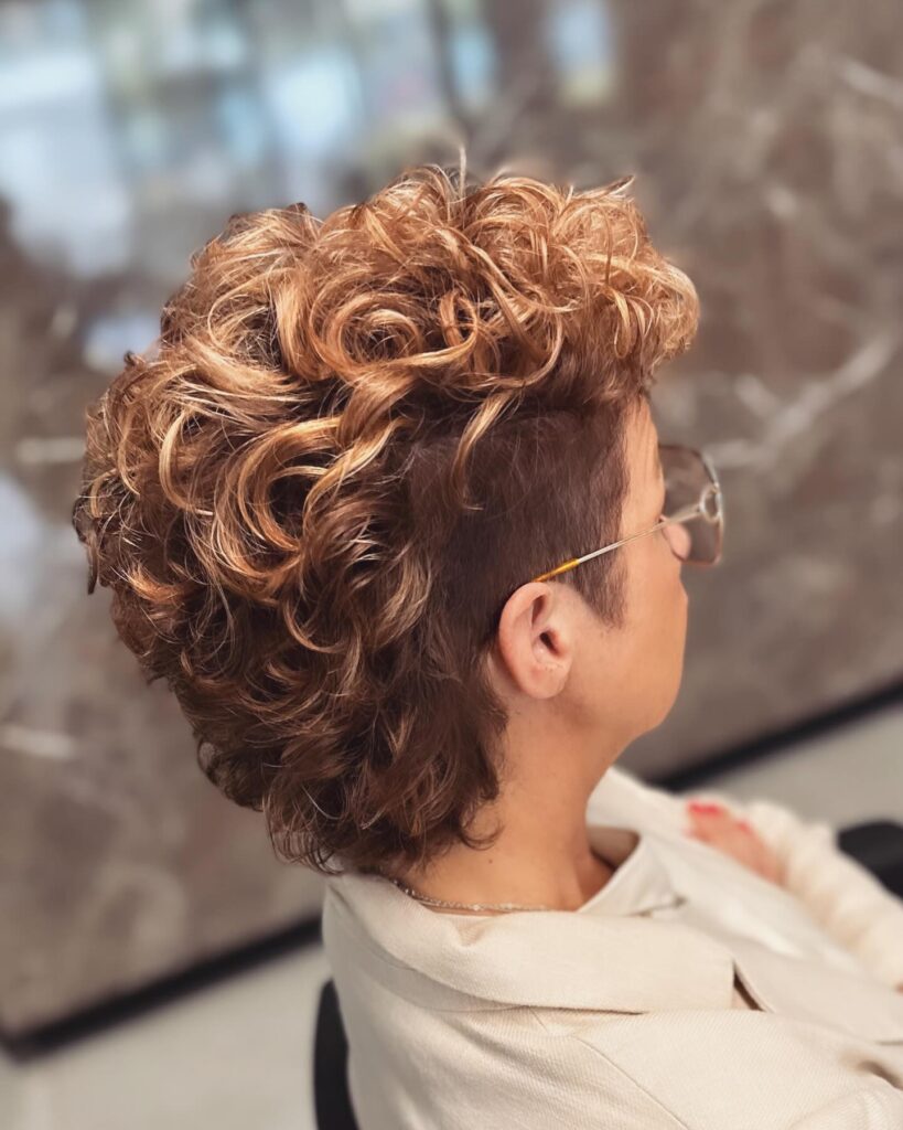 A side and back view of a woman with a short curly hairstyle featuring buzzed sides and voluminous honey-blonde ringlets on top.