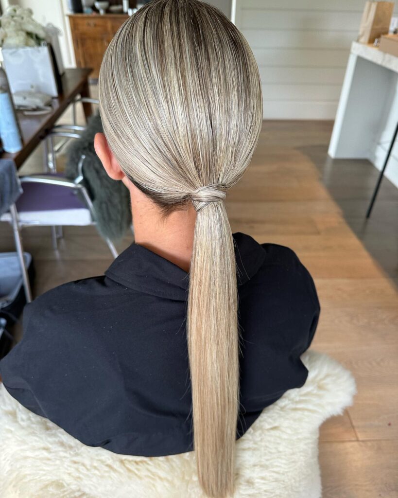 A woman from behind with long, straight ash-blonde hair styled into a sleek low ponytail with a hair wrap around the base.
