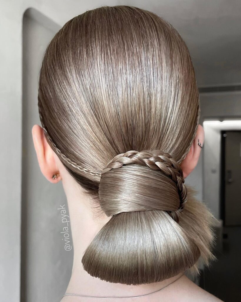 A woman from behind with ash blonde hair styled in a sleek low chignon with a decorative braid wrap and side accent braids.