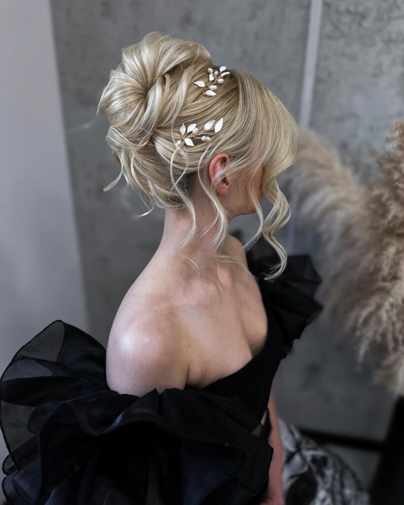 A high-angled photograph captures a woman with blonde hair styled into a complex, textured French twist adorned with delicate white enamel leaves and pearl hair accessories.
