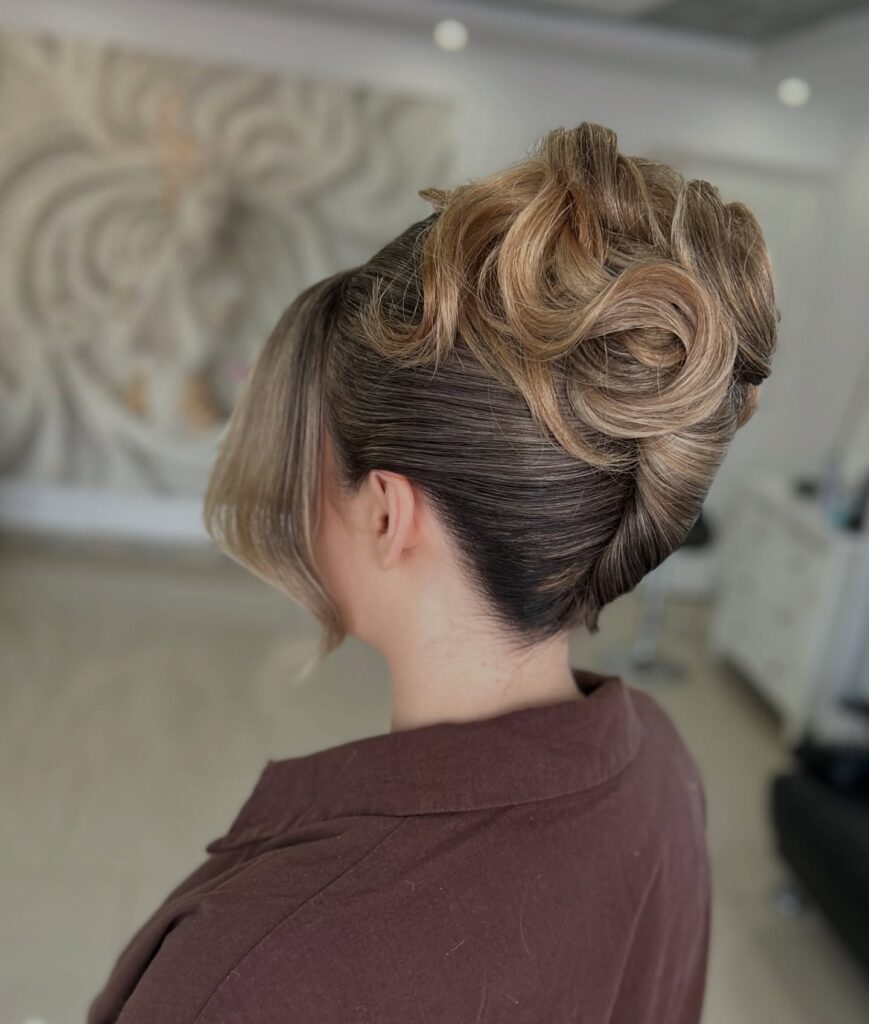 A woman with brunette hair and caramel highlights displays a sculpted French twist featuring intricate circular swirls on top and a sleek side-swept fringe.