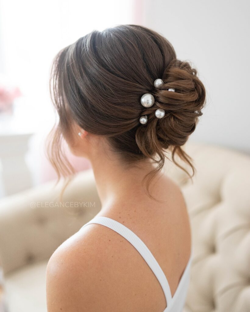 A woman with dark brunette hair wears a low and soft French twist decorated with several large white pearl hairpins.