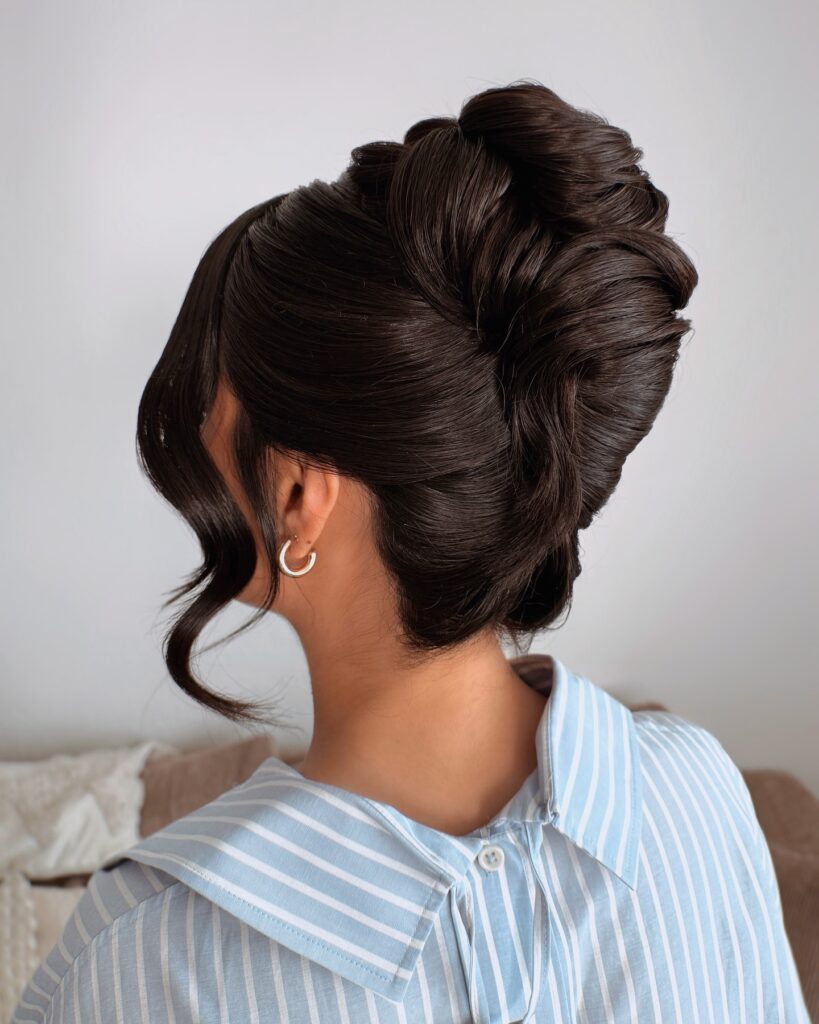 A woman with shiny dark brunette hair displays a high and perfectly sculpted vertical French twist with a single long curl framing her face.