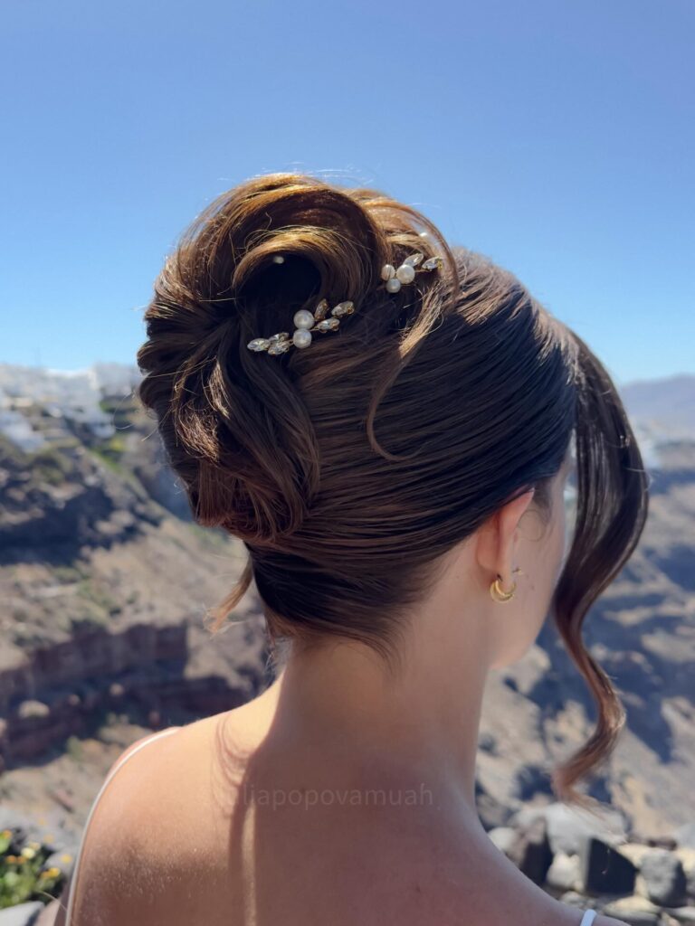 A woman with chestnut brunette hair showcases a textured French twist accented with delicate pearl and jewel hairpins while standing against a scenic coastal background.