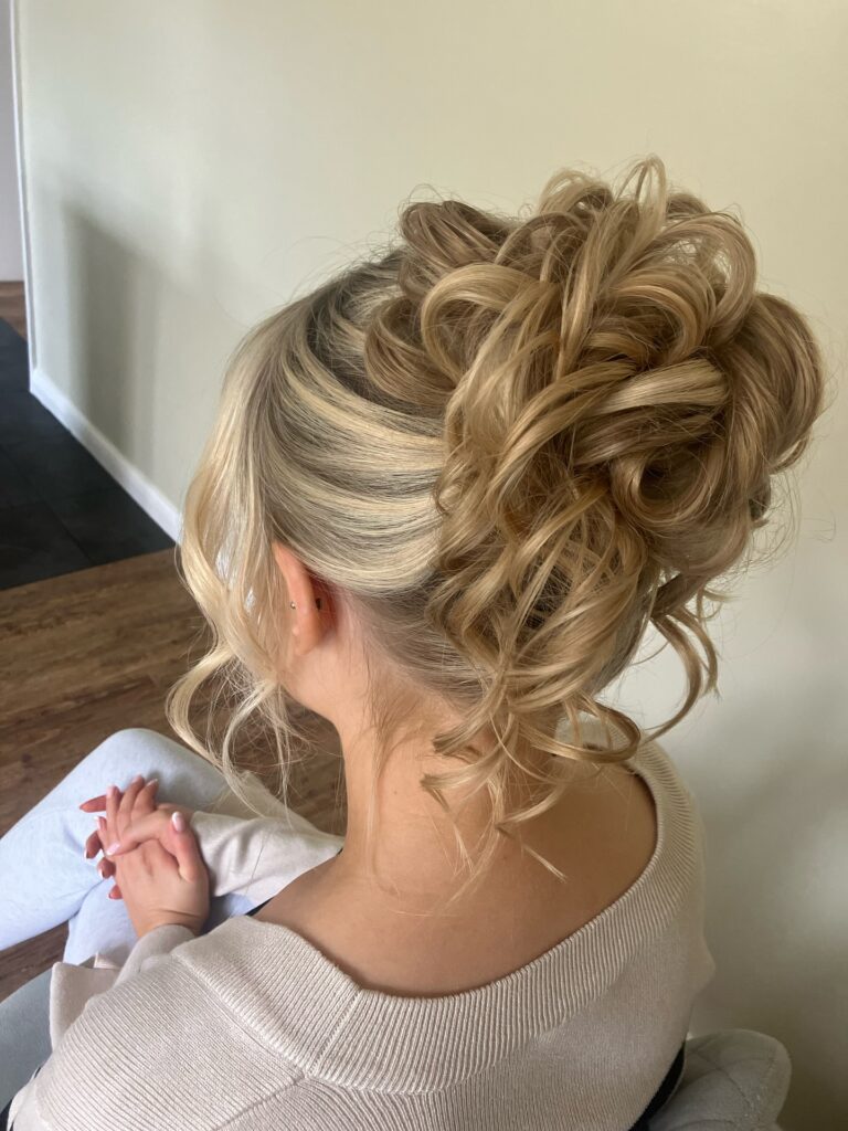 A woman with champagne blonde hair displays a French twist that transitions into a voluminous cluster of bouncy curls at the crown.