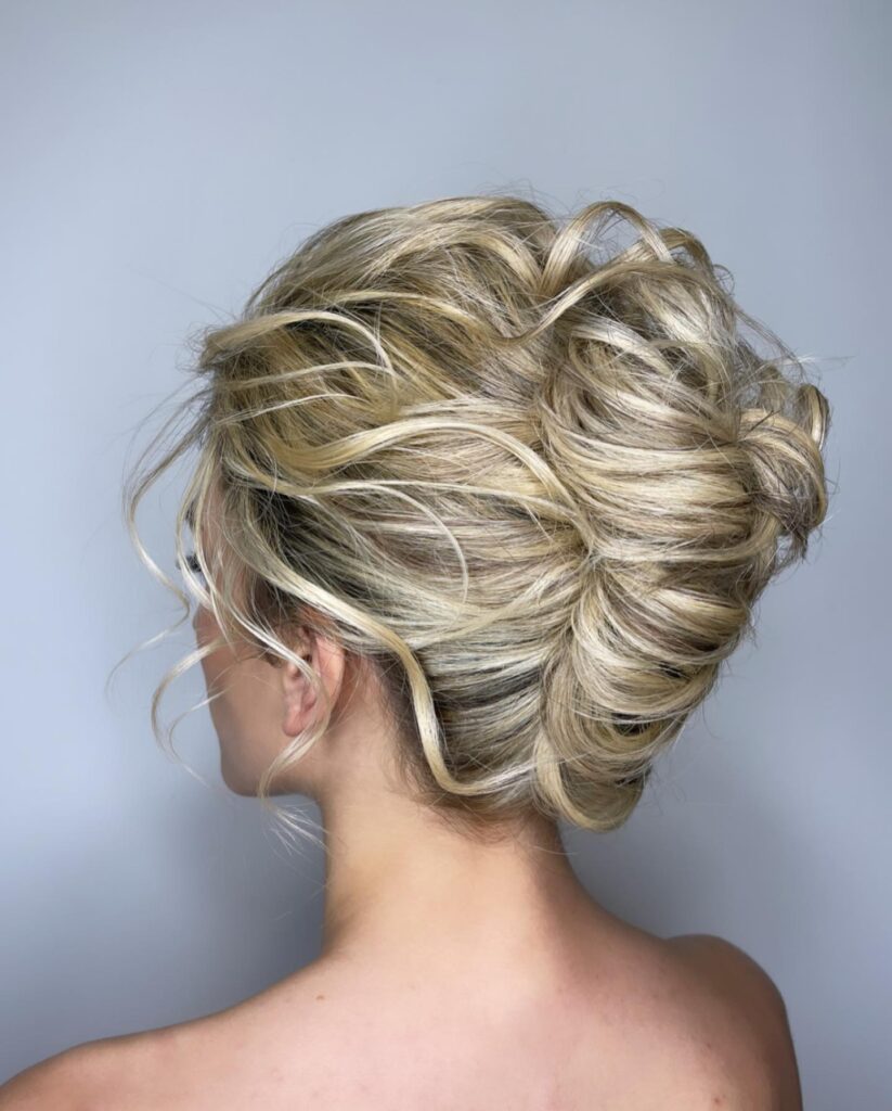 A woman with ash-blonde hair features a very loose and messy French twist with heavy texture and wavy face-framing pieces.