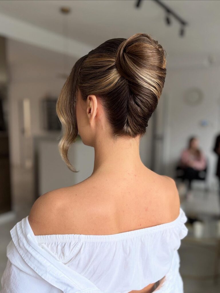 A woman with brunette and caramel-highlighted hair displays a perfectly sleek and smooth vertical French twist with a single wavy strand framing her face.