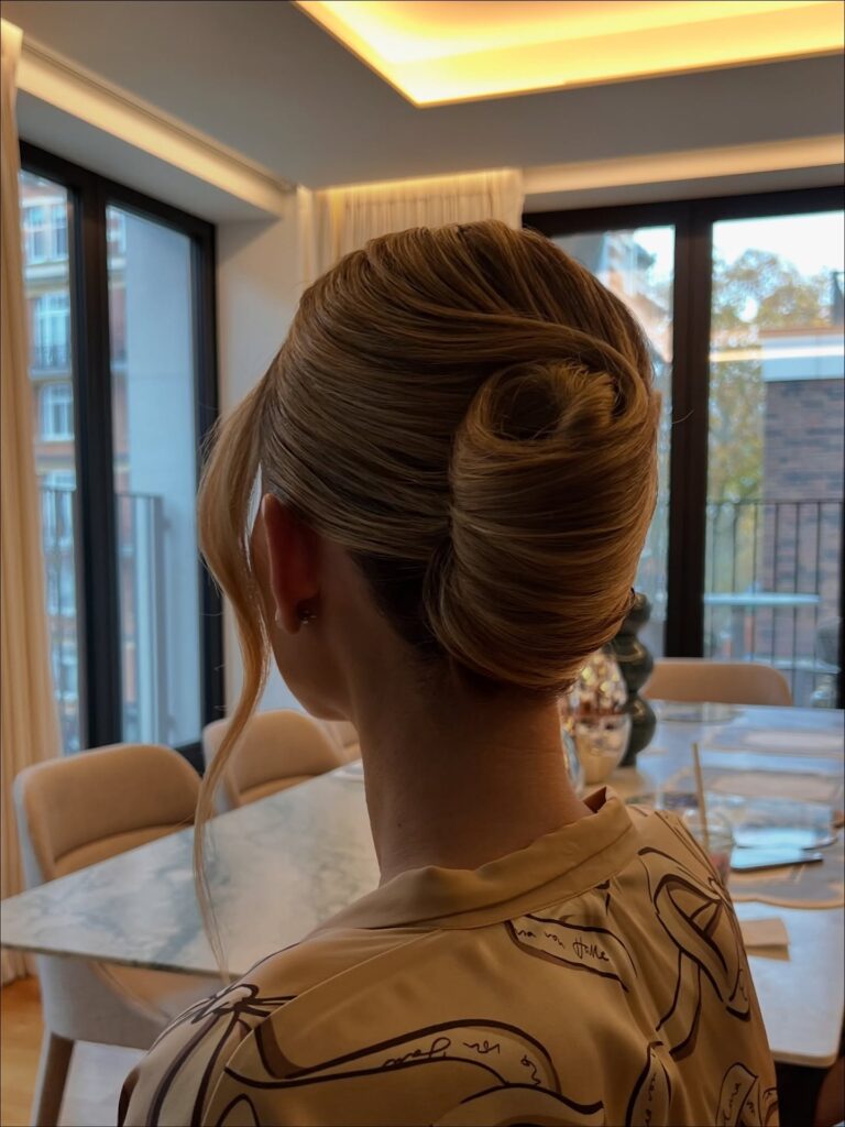 A woman with honey blonde hair shows off a sleek and tightly tucked French twist featuring a large decorative hair swirl at the top and a smooth face-framing strand.