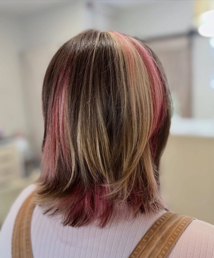 A back view of a woman with a medium-length brown and blonde hairstyle featuring vibrant pink highlights peeping through the layers.