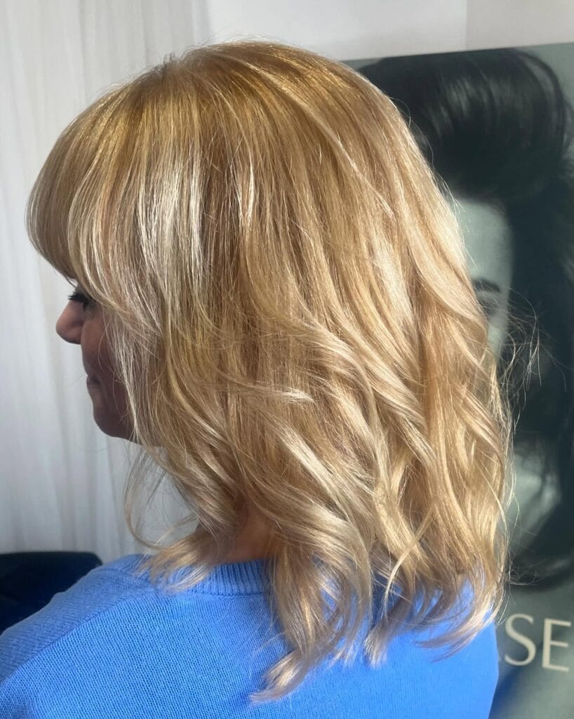 A side view of a woman with a medium-length champagne blonde hairstyle featuring a full fringe and soft waves.