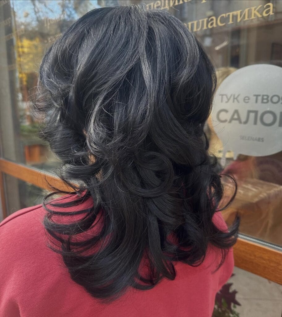 A back view of a woman with medium-length, jet-black hair styled in thick cascading layers and large, defined curls.