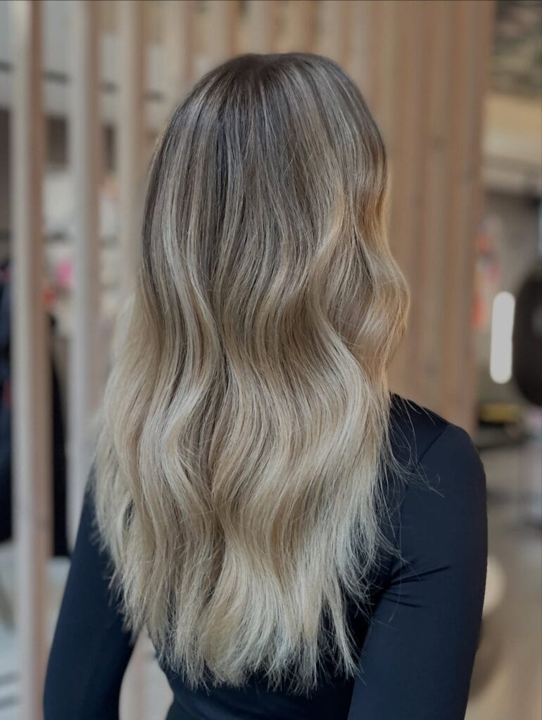A back view of a woman with medium-length hair featuring a mushroom brown to ash blonde ombre, styled in soft waves.