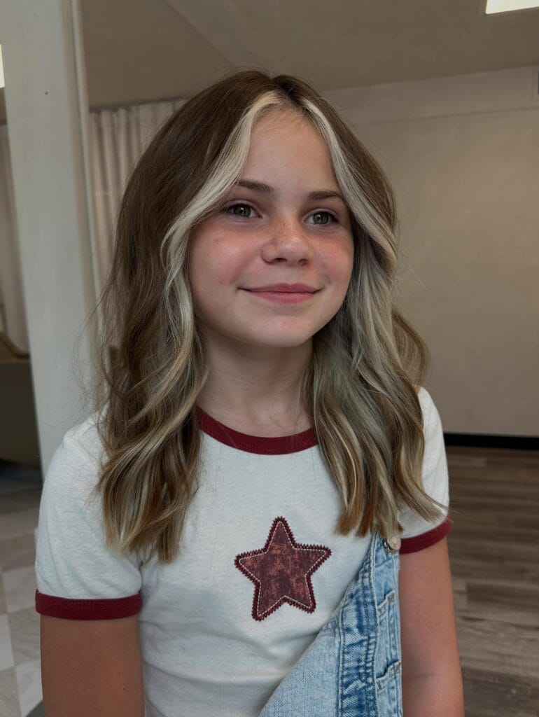 A young girl with medium-length brown hair featuring bold blonde, face-framing highlights and soft waves.