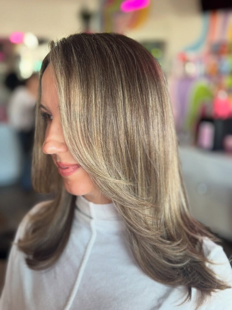 A side profile of a woman with medium-length ash-blonde hair featuring long, sweeping layers and a silky-smooth finish.