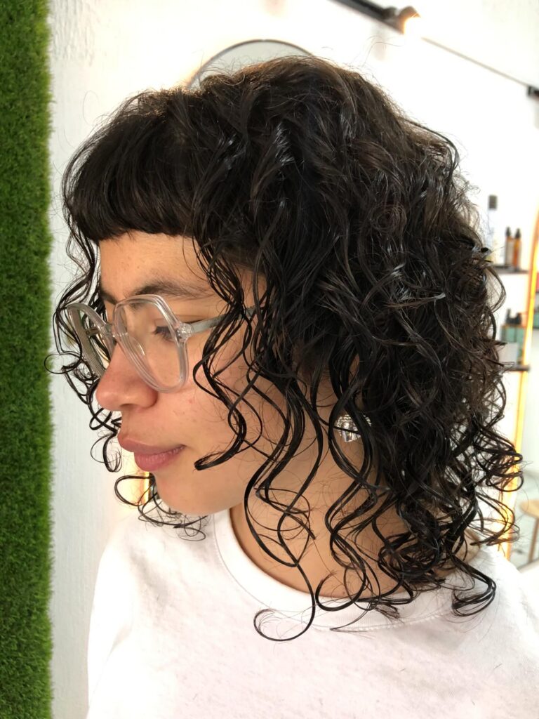 A woman in profile with a dark brown curly shaggy haircut and short straight micro-bangs.