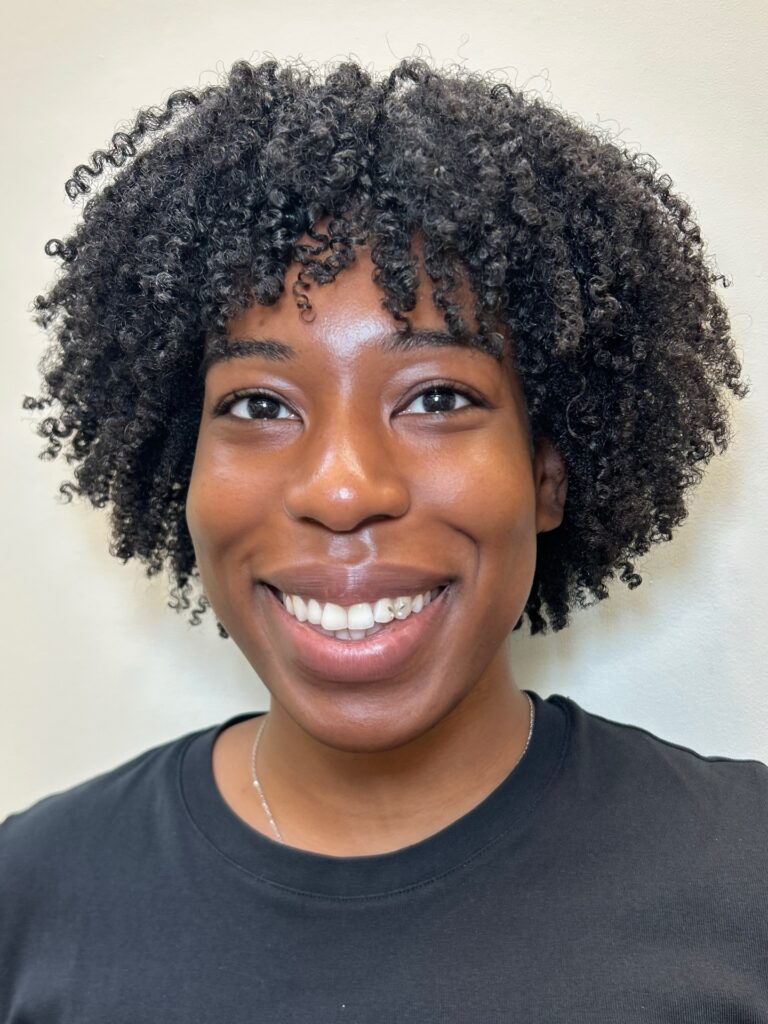 A smiling woman with a rounded chin-length haircut featuring tight black coils and a soft curly fringe.