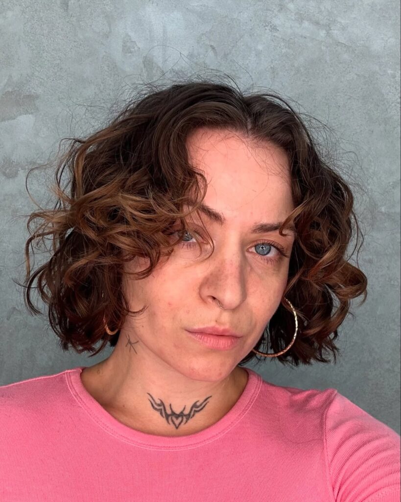 A woman with a center-parted curly bob in a warm brown shade with caramel highlights.