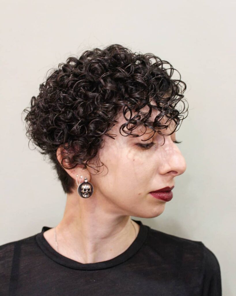 A woman in profile wearing a dark espresso tapered pixie cut with tight glossy curls and a wispy fringe.