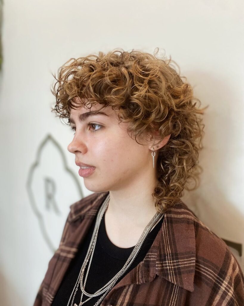 A woman in profile wearing a golden blonde curly mullet with shaggy layers and a full fringe.