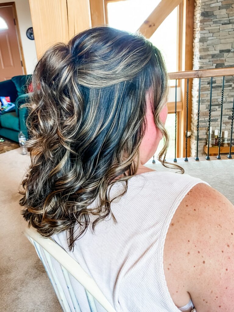 A side view of a woman with medium-length dark hair and blonde highlights styled in a half-up look with bouncy curls.