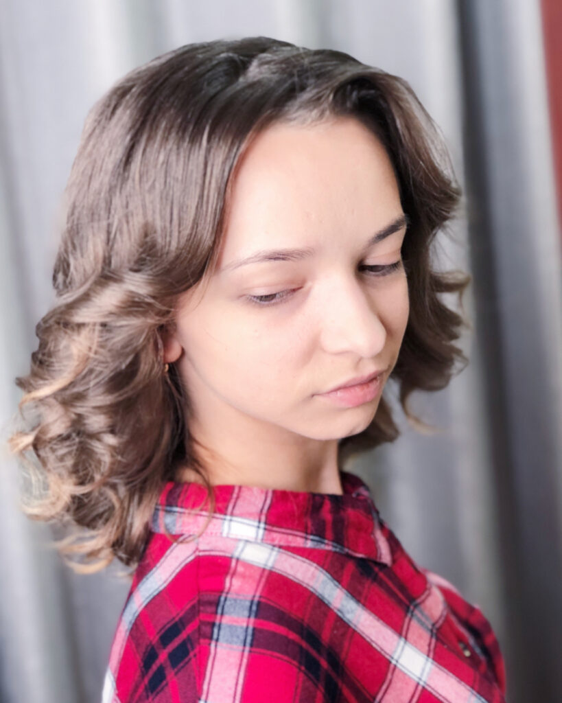 A woman with shoulder-length brunette hair styled in bouncy curls with caramel highlights at the tips.