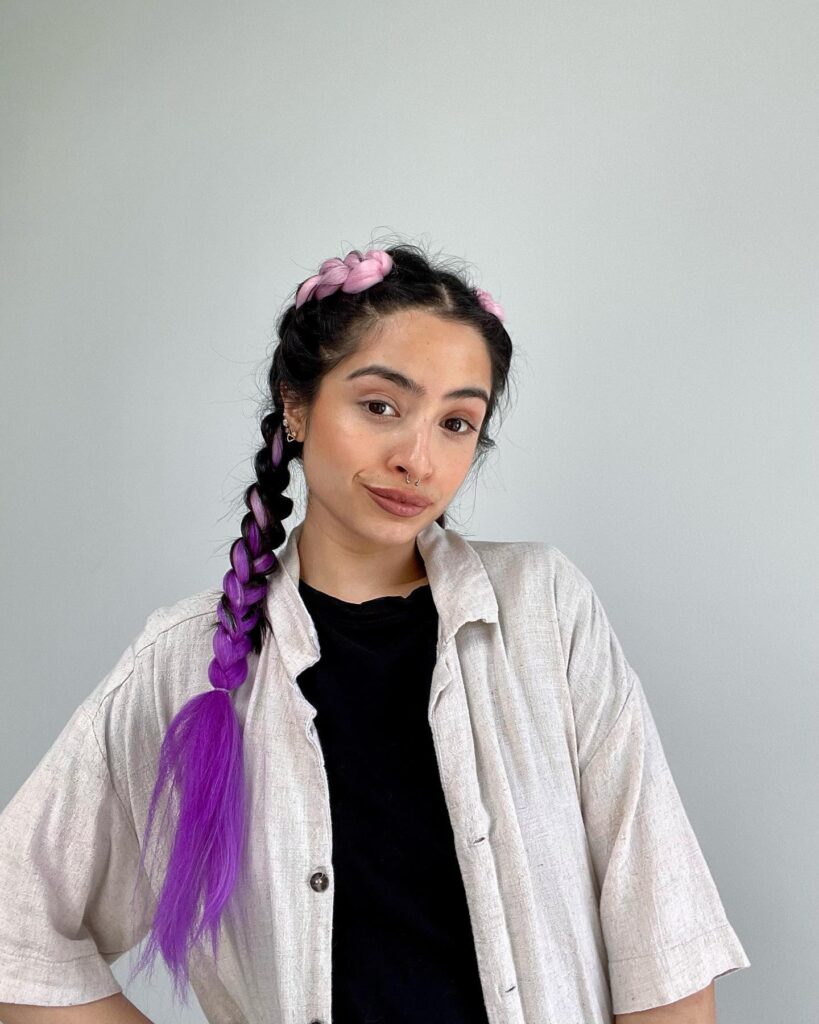 A woman with dark hair styled in a thick Dutch braid featuring light pink and bright purple extensions that transition into a long purple ponytail.