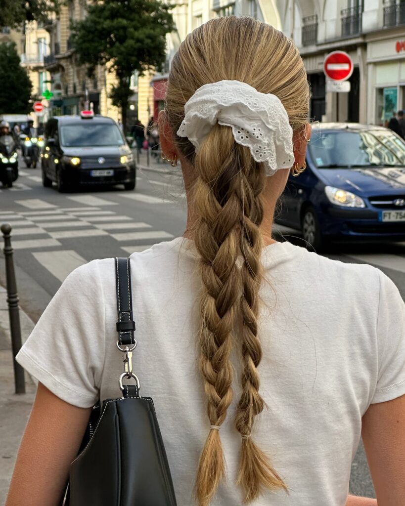 A woman from behind walking down a city street with her blonde hair in a low ponytail secured by a white eyelet lace scrunchie and split into two braids.