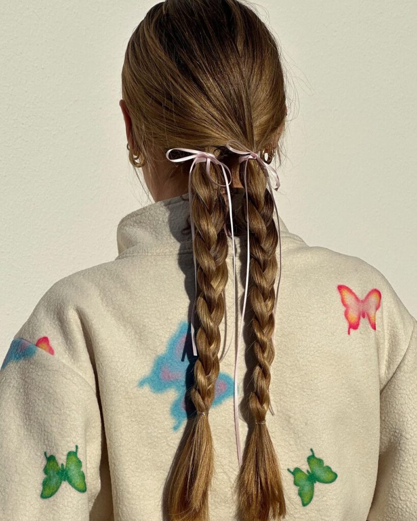 A woman from behind with long caramel brown hair styled into two neat pigtail braids, each featuring a delicate, thin pink ribbon tied into a sweet bow at the top, wearing a fleece jacket with colorful butterfly patches.
