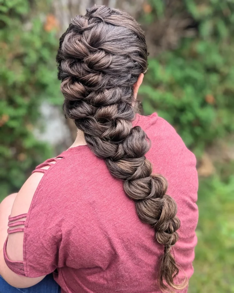 A woman viewed from behind with extremely long and thick brunette hair with subtle highlights styled into a voluminous, loose low Dutch braid.