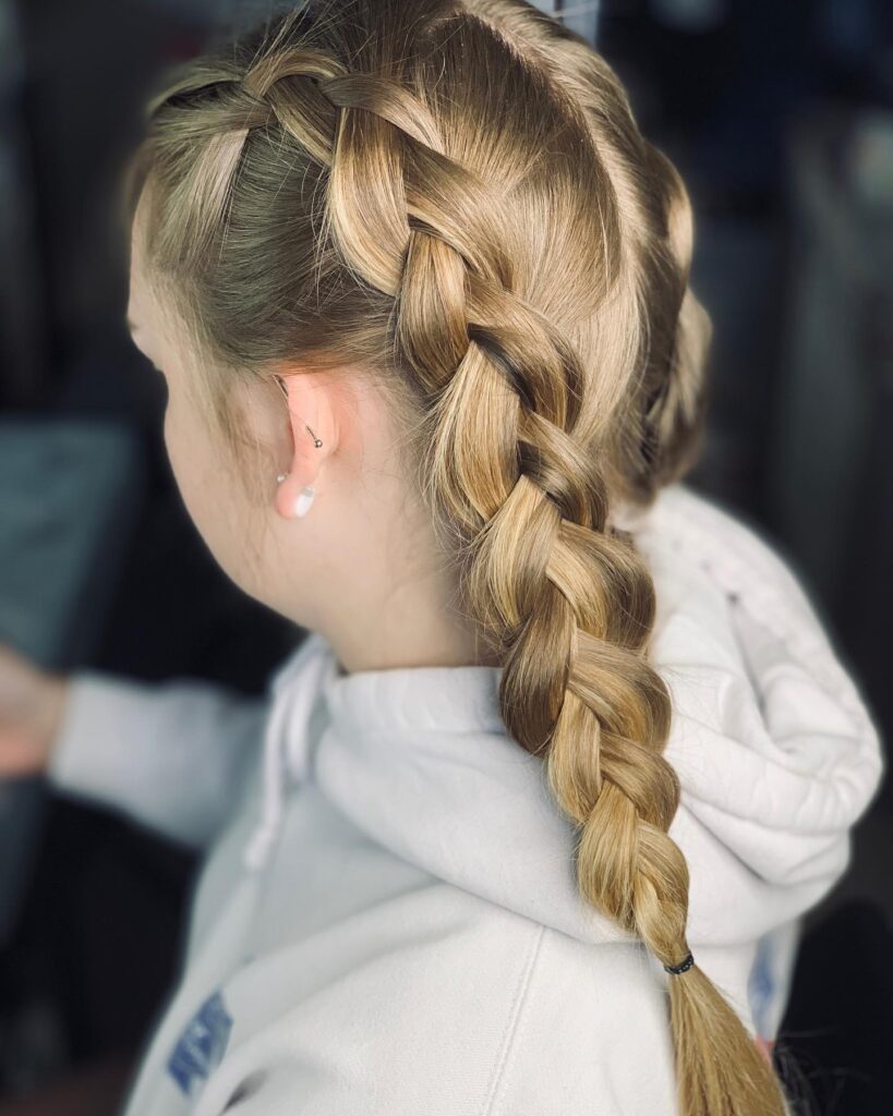 A young woman with long blonde hair viewed from the back and side, styled into two neat and sporty French braided pigtails, wearing a white hoodie.