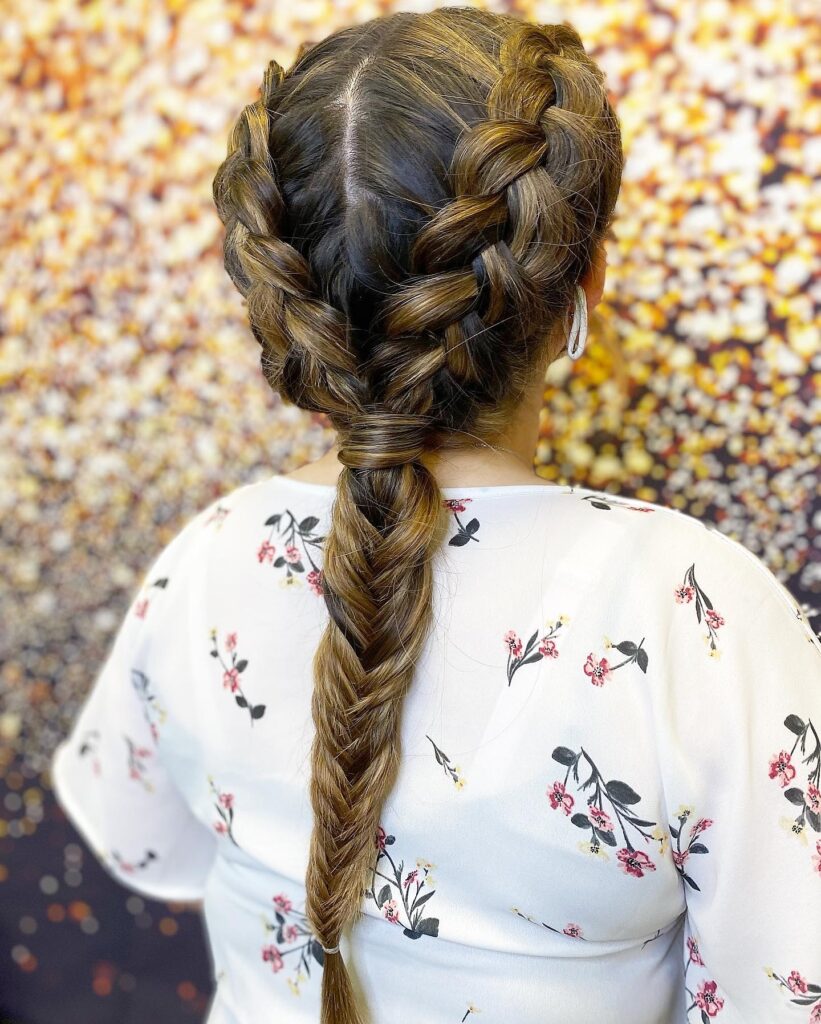 A woman with highlighted brown hair from behind showing two Dutch braids that transition into a single long fishtail braid.