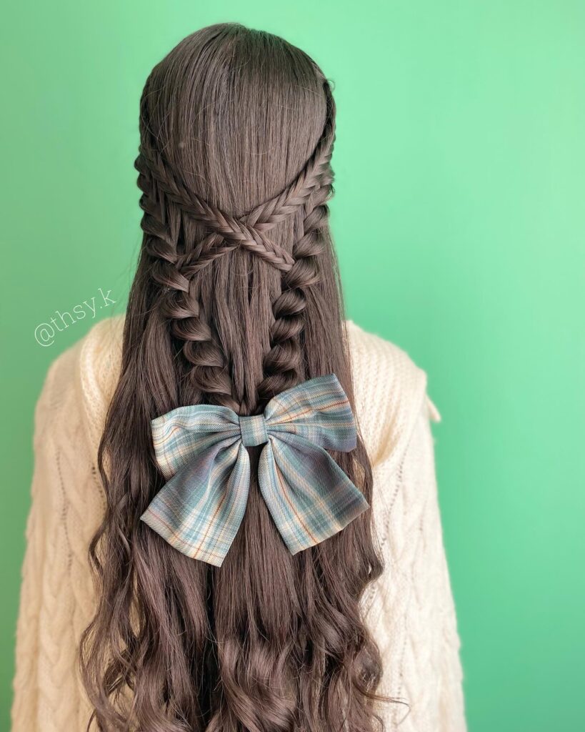 A woman with long wavy brown hair from behind featuring a complex crisscross braided half up style finished with a large blue plaid bow.