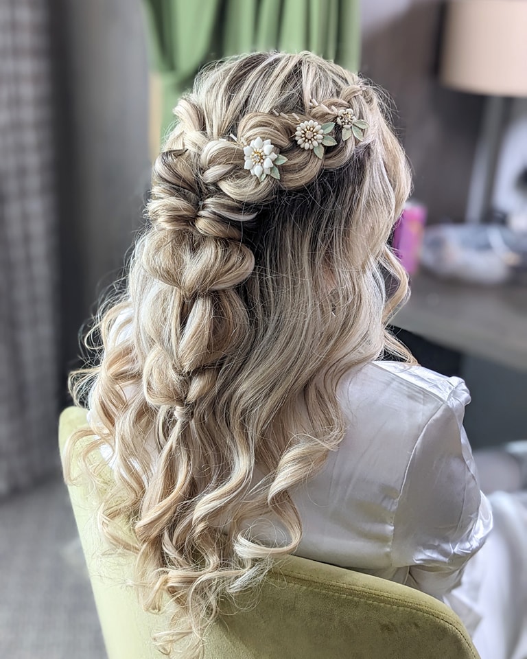 Long blonde wavy hair styled in a half-up braid with floral accessories woven into the braid.