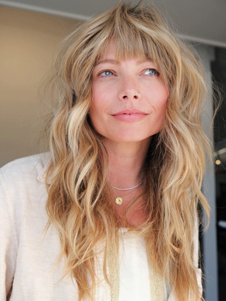 A woman with long wavy sunkissed blonde hair and soft piecey bangs framing her face.