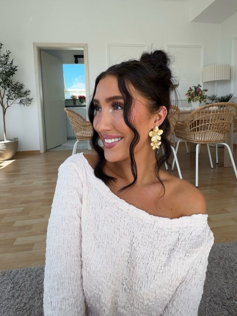 A woman smiling with her dark hair styled in a high messy bun and long wavy strands framing her face.