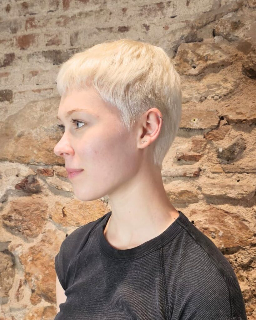 A woman with a very short platinum blonde pixie cut featuring a micro fringe and tapered sides.