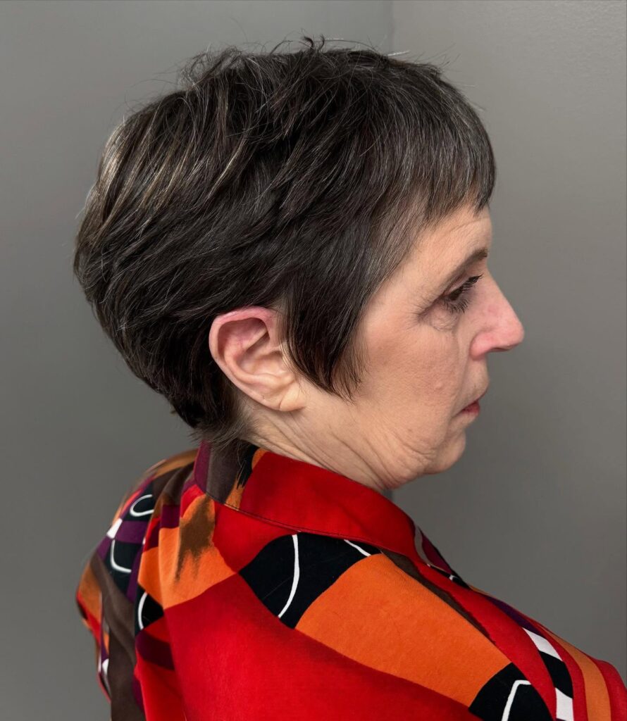 A side profile of a woman with a salt and pepper pixie cut styled with textured layers and a short wispy fringe.