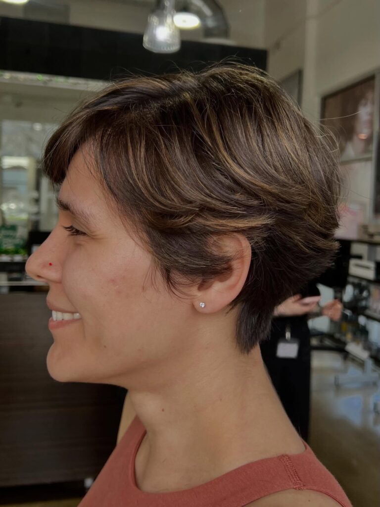 A woman with a layered brunette bixie cut featuring golden highlights and soft wispy layers flicked out at the back.