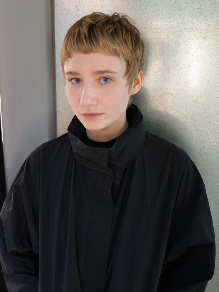 A young woman with a short golden-blonde pixie cut that has fine layers and a piecey micro fringe.