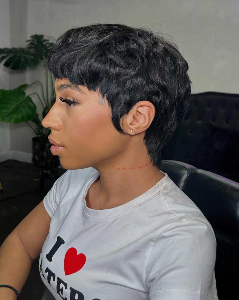 A side profile photograph of a woman with short, textured, wavy dark black hair in a chic pixie cut featuring defined layers and stylized sideburns.