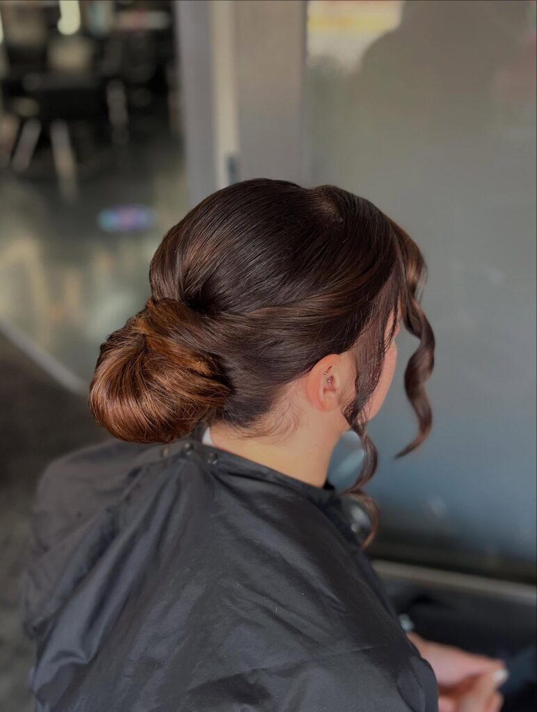 A woman with glossy dark brown hair styled in a sleek low bun with two curly face framing strands.
