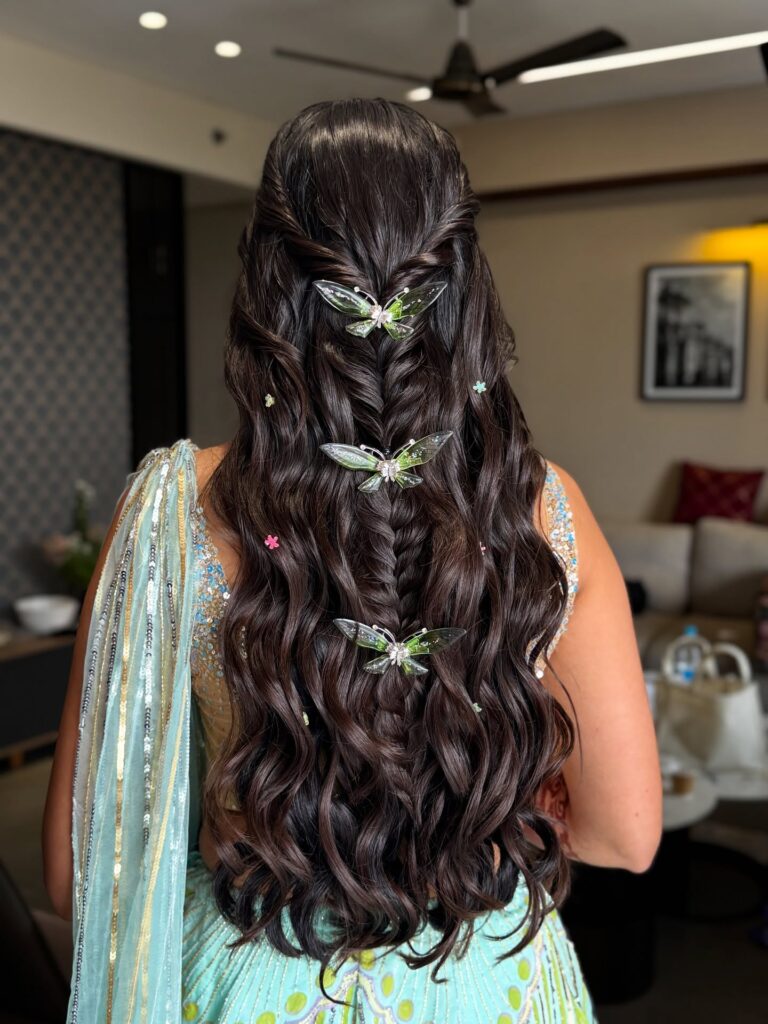 A woman with long dark wavy hair styled in a twisted half updo and fishtail braid adorned with three green butterfly clips and small floral pins.