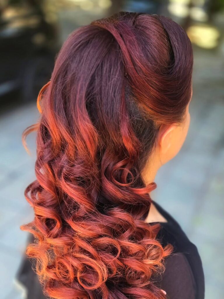 A woman with long wavy hair in a gradient of burgundy and copper styled in a polished half updo with defined ringlet curls.