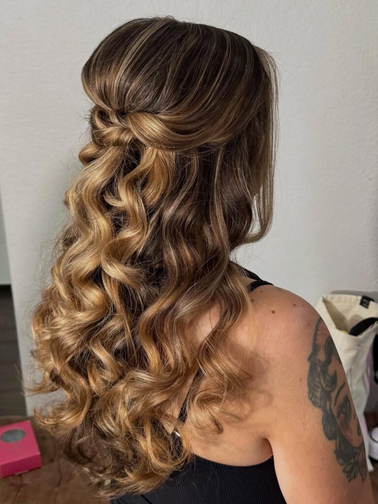 A woman with long wavy hair in a honey blonde balayage styled in a smooth half updo with voluminous defined curls.