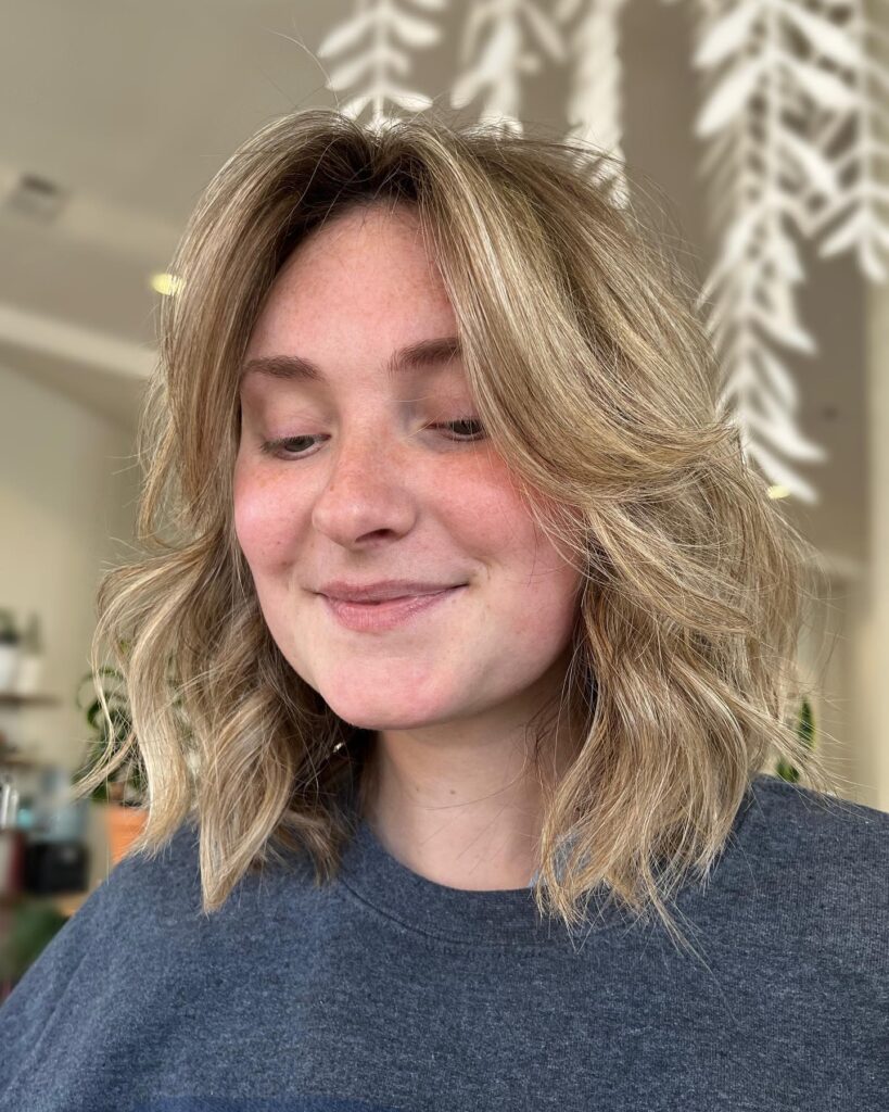 A woman with a shoulder length textured blonde lob featuring sun kissed highlights and soft curtain bangs.
