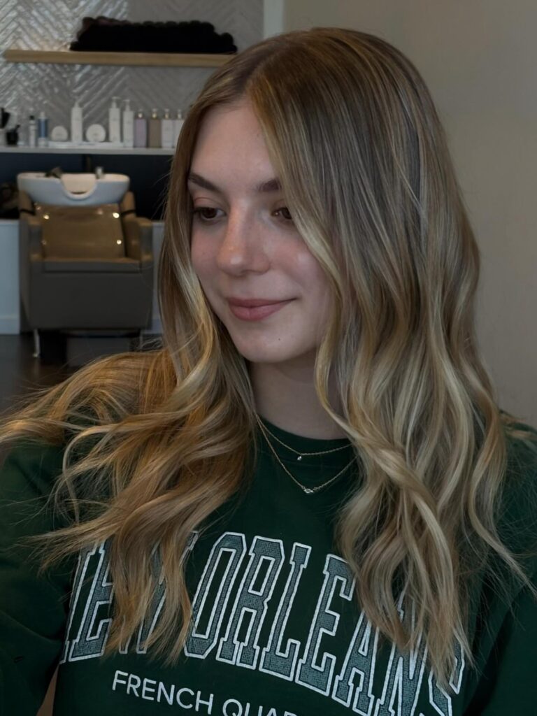 A woman with long wavy brown hair featuring warm honey caramel balayage highlights and a middle part.
